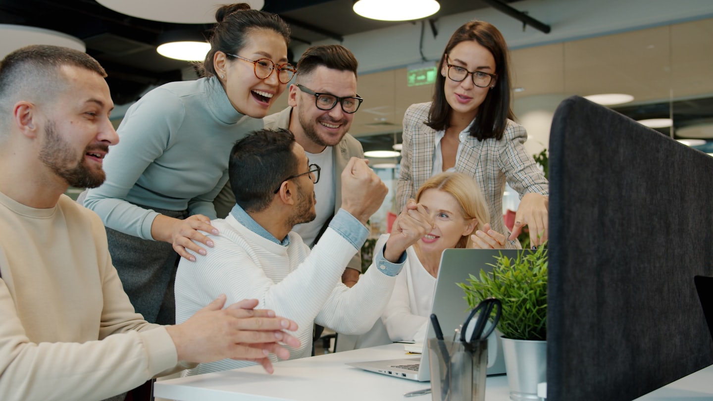 Un team eterogeneo festeggia il successo attorno a un laptop in un ufficio moderno.